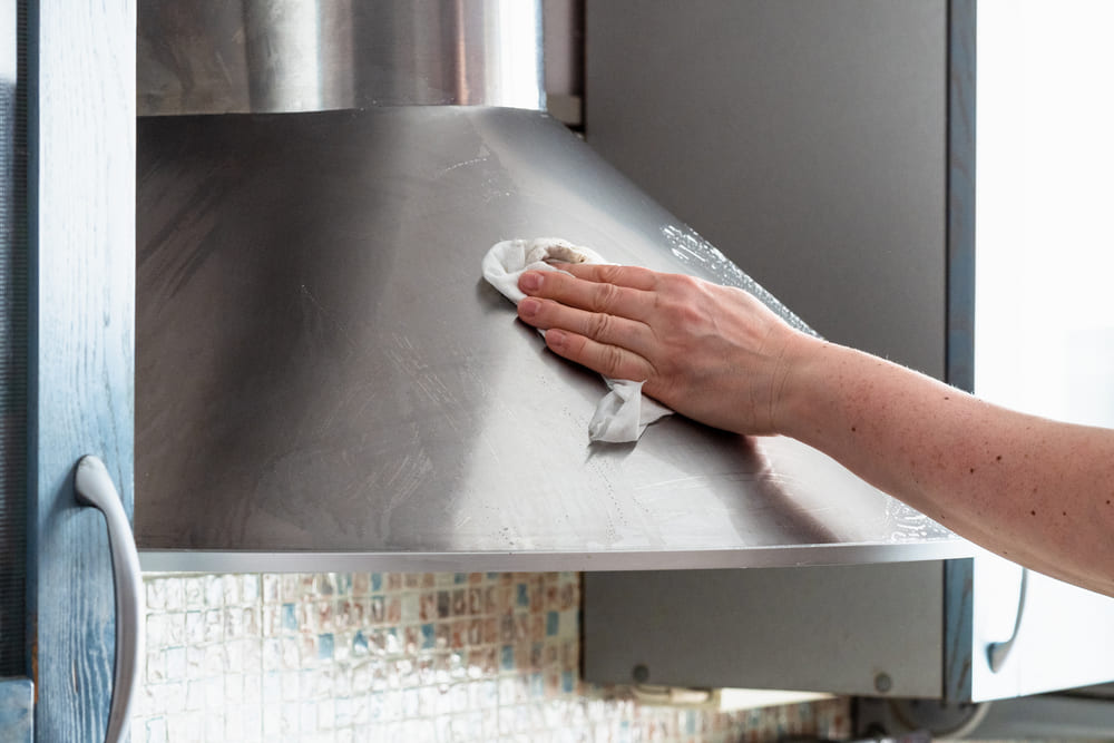 A hand with a rag, gently cleaning a range hood made of stainless steel A hand with a rag, gently cleaning a range hood made of stainless steel.