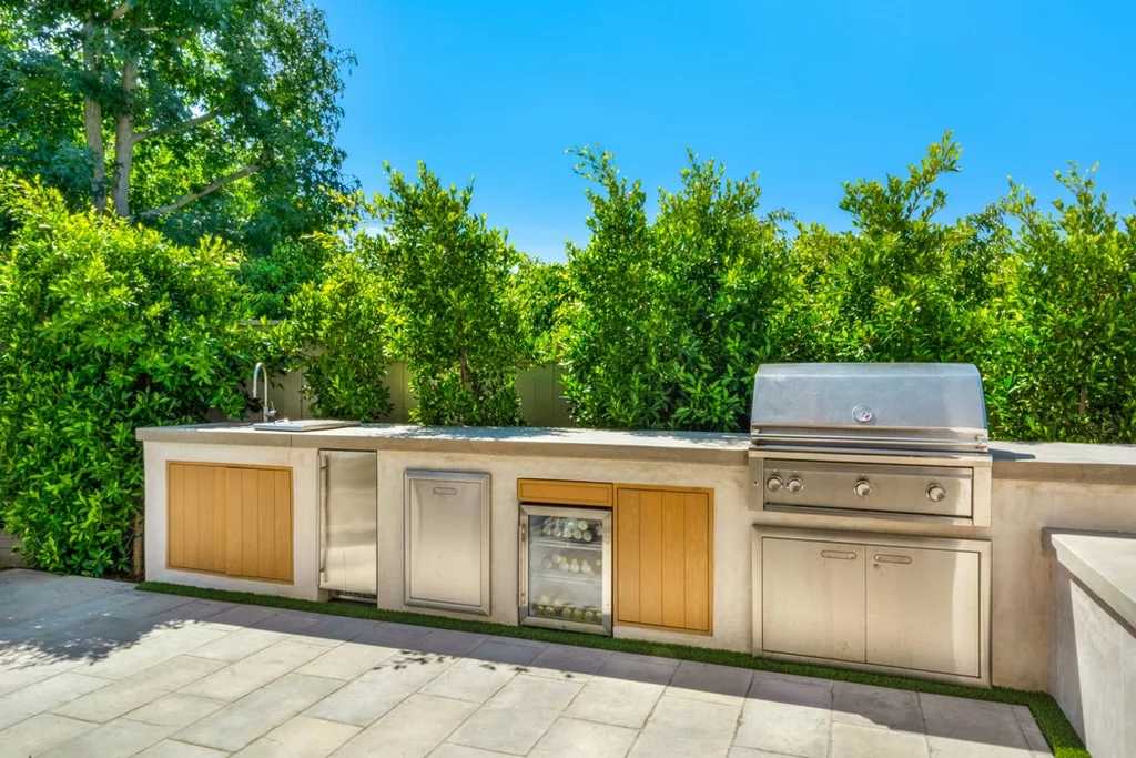 Outdoor kitchen with built-in grill, storage, and outdoor refrigerator drawers surrounded by greenery under clear sky.